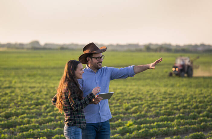 Two farmers man and woman with tablet standing in soybean field in front of tractor at sunset