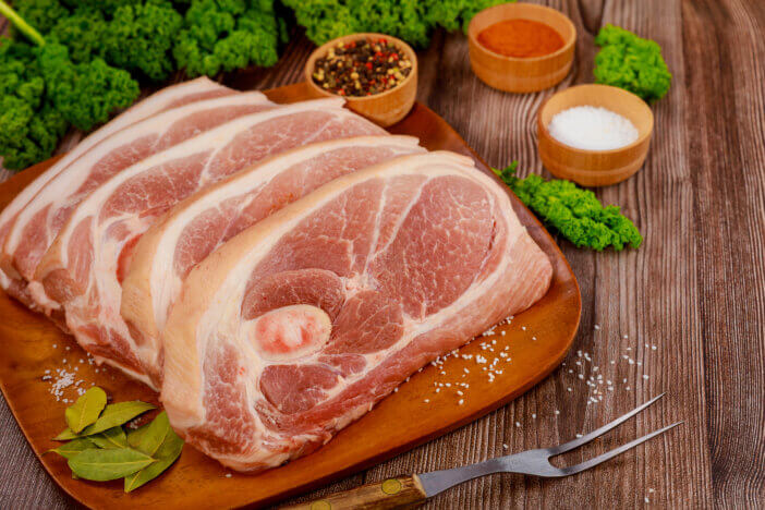 Sliced raw pork shoulder with spices and fork on wooden board.
