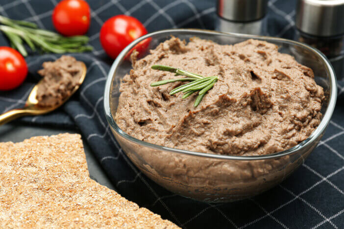 Tasty liver pate with rosemary in glass bowl and crispy crackers on table, closeup