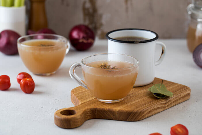 Homemade bone broth in mugs with spices on a wooden stick with bay leaf, tomatoes and onions. High quality photo