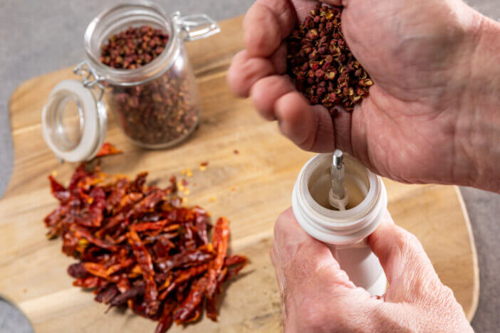 A hand fills a pepper mill with dried pepper in a kitchen over a wooden board.