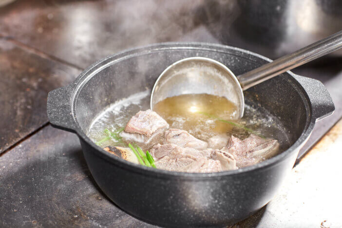 chef cook adds the ingredients. traditional beef broth with vegetable, bones and ingredients in pot, cooking recipe. Soup in a cooking pot with ladle on dark stone background.