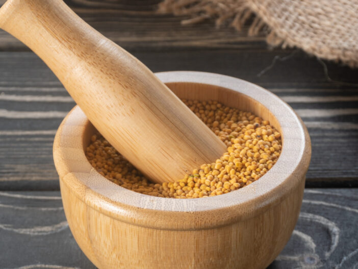 Mustard seeds in a mortar and ground mustard seeds in the spoon on brown wooden table