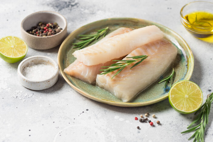 Ceramic plate with fresh raw cod fish, aromatic herbs, spices, lime and olive oil on kitchen table. copy space