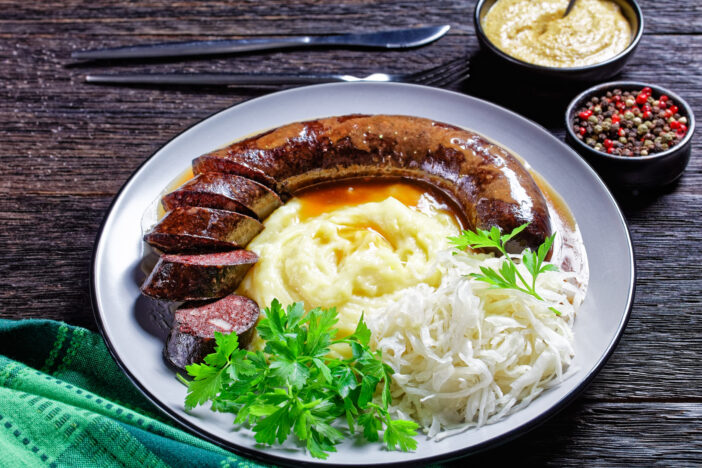 German food: blutwurst or blood sausage served on a plate with sauerkraut, mashed potato parsley, mustard, and peppercorns on a dark wooden table, top view, close-up