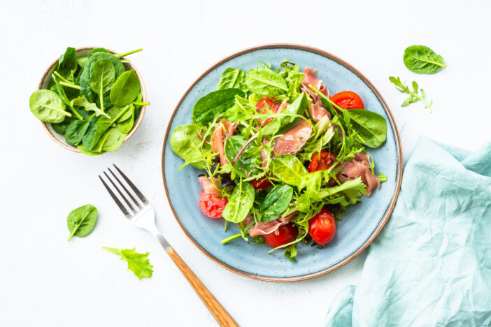 Green salad with fresh leaves, tomatoes and jamon at white table. Top view with copy space.