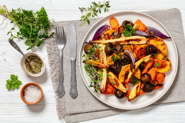 baked winter vegetables and mushrooms, potato, winter squash, red onions, carrots, parsnip on plate on white wooden table with cutlery and fresh herbs, horizontal view from above, flat lay