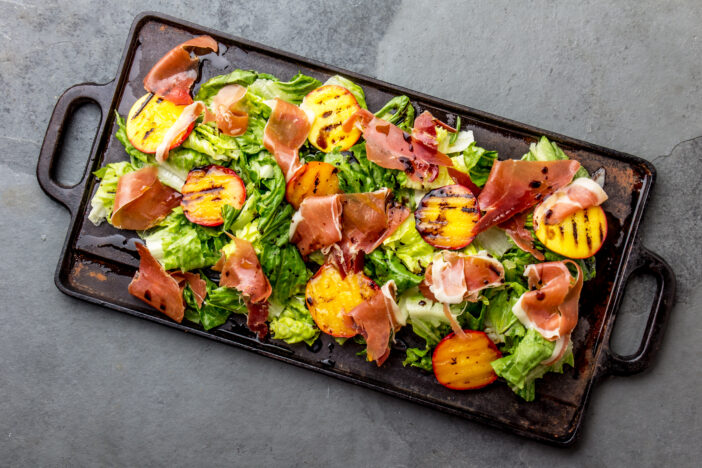 Lettuce, ham serrano and grilled peaches salad served on black board. Top view.