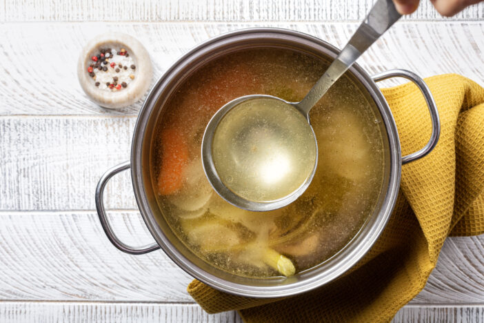 Cooking beef broth stock in a saucepan with a ladle , made using meat, bones, vegetables, carrot, onin, celery, and seasoning.