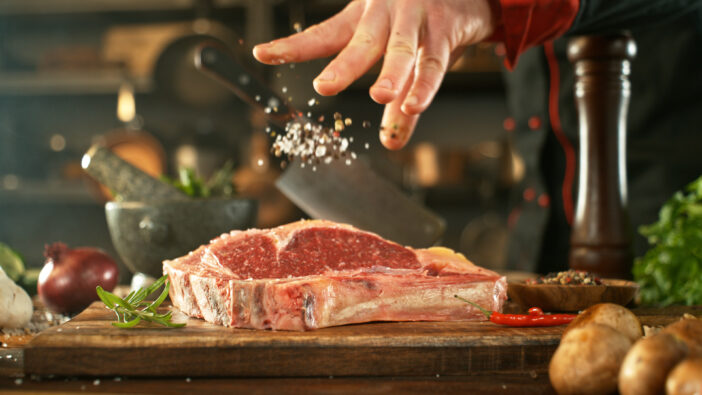 Close-up of falling salt on tasty beef steak in kitchen. Chef in background.