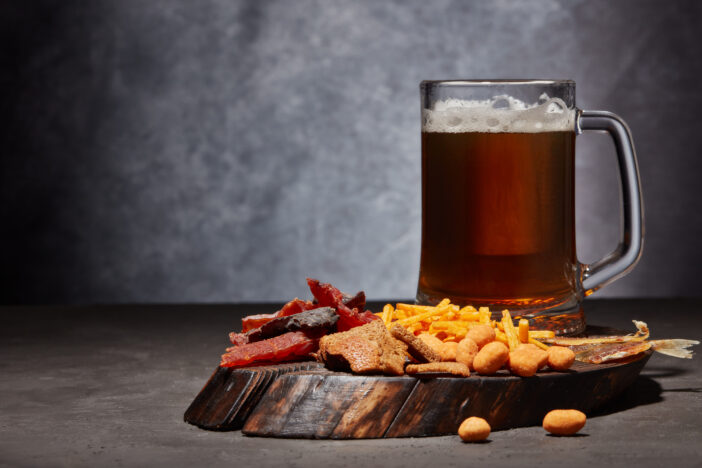 Glass mug with beer and set of various kind snacks: jerky, potato chips, salted nuts, stockfish, bread crackers on brown wooden tray on dark gray textured  background. Snacks for beer.