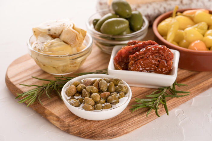 Wooden board with selection of antipasti served on white table background - pickled artichokes, capers, baby peppers, sun dried tomatoes and olives with rosemary