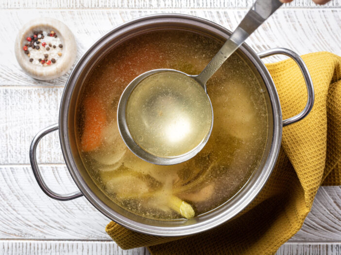 Cooking beef broth stock in a saucepan with a ladle , made using meat, bones, vegetables, carrot, onin, celery, and seasoning.