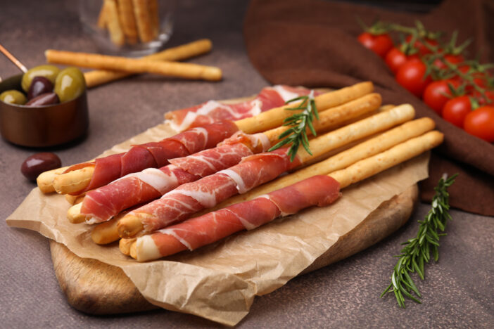 Delicious grissini sticks with prosciutto and ingredients on brown textured table