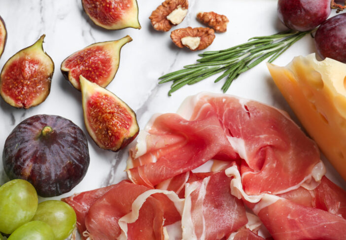 Delicious ripe figs, prosciutto and cheeses served on white board, flat lay