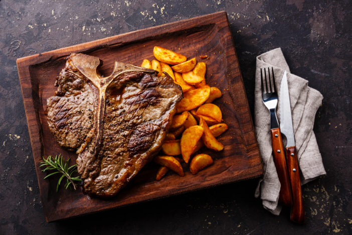 Medium rare Grilled T-Bone Steak with potato wedges on serving board block on dark background