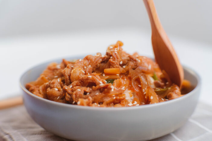 Korean food - Hands holding a spoon eating stir-fried kimchi and kurobuta pork with steamed rice in a bowl. Korean homemade food for lunch