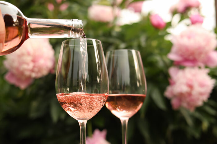 Pouring rose wine from bottle into glass against beautiful peonies, closeup