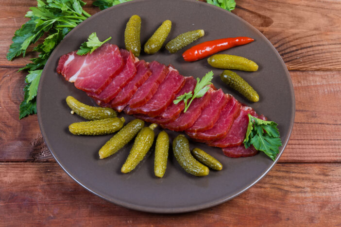 Slices of dried-smoked pork neck among the pickled gherkins on the brown dish on the rustic table close-up