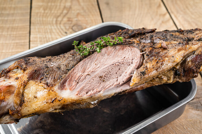 Whole oven roasted goat leg meat with thyme in a steel tray. Wooden background. Top view.