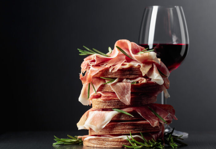 Spanish jamon or Italian prosciutto with bread, rosemary, and red wine on a black wooden table.