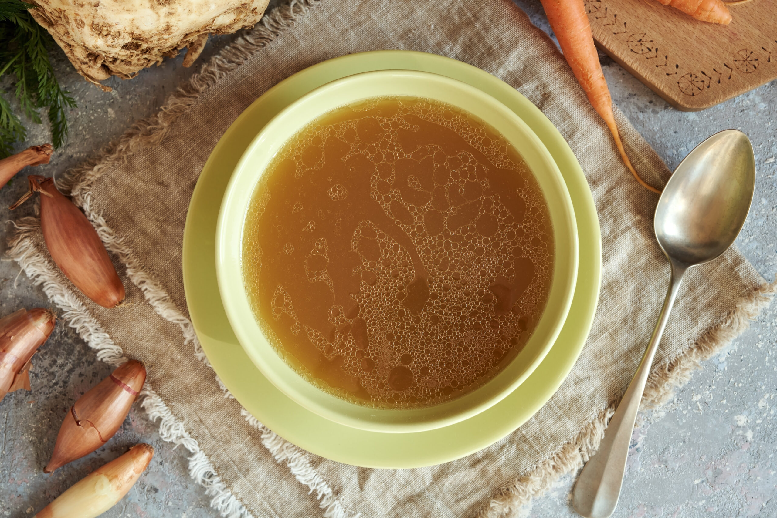 Beef bone broth or soup in a green soup bowl with fresh vegetables, top view