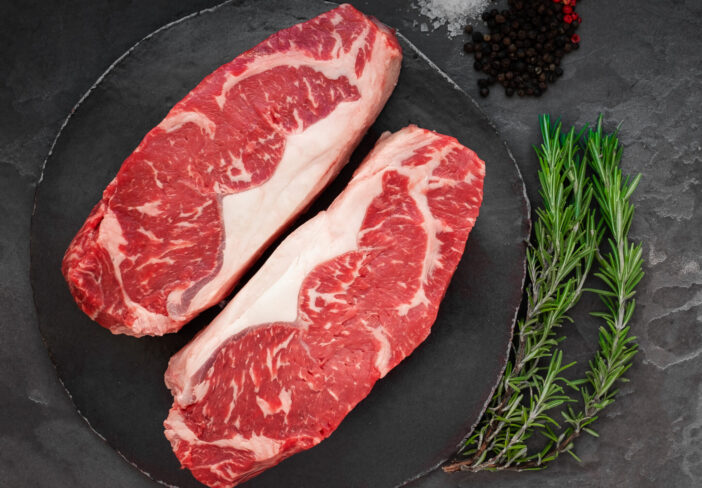 Raw striploin beef steak with rosemary, salt and pepper on round slate on stone table. Top view