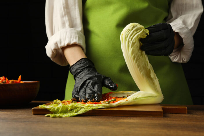 Woman preparing spicy cabbage kimchi at wooden table against dark background, closeup