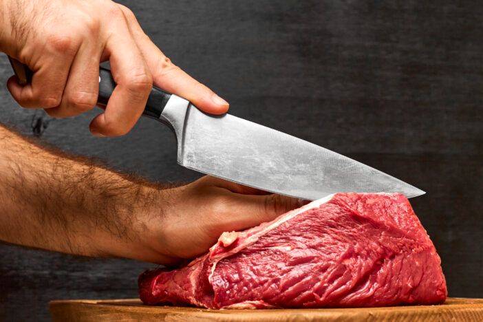 Closeup man hand cutting a beaf meat with a knife on a black background. Concept of healthy protein meal