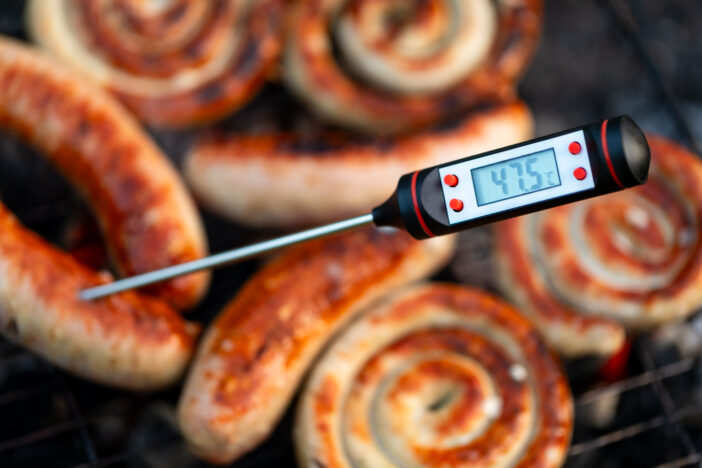 Close-up shot of a digital meat thermometer displaying temperature below the safe cooking threshold for grilled sausages - signaling a potential health risk due to insufficient heat to kill harmful germs