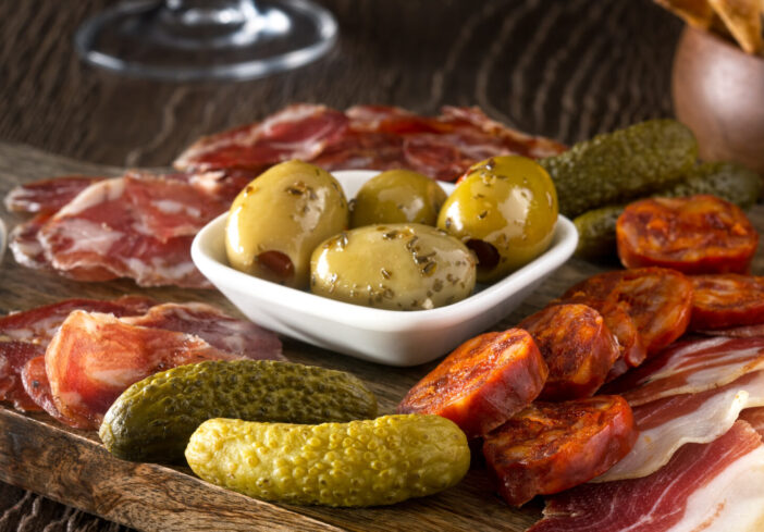 A delicious charcuterie assortment of meat, olives, gherkins, and pickled peppers with breadsticks on a wooden background.