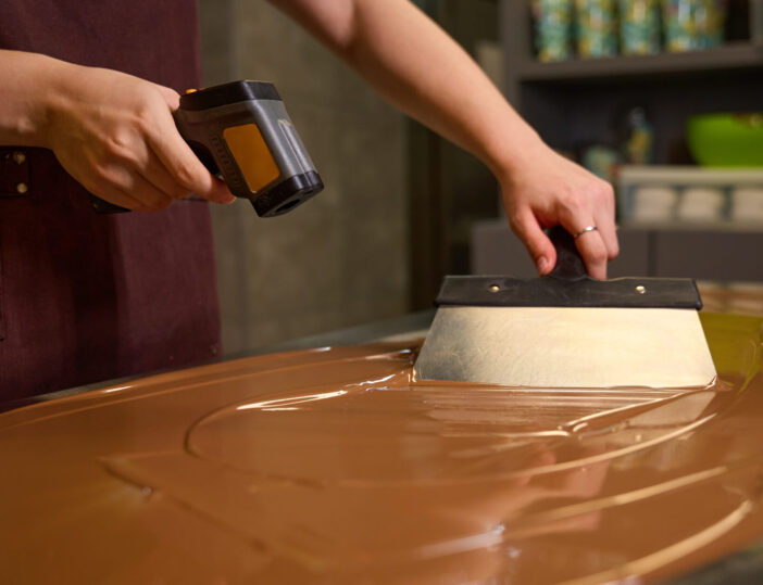 Cropped photo of chocolatier stirring melted chocolate on worktop with spatula while checking its temperature with infrared thermometer