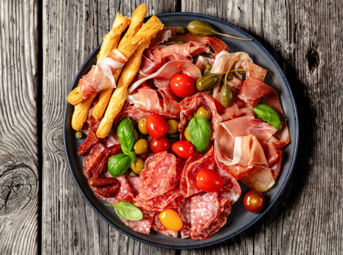 italian antipasto platter of salami slices, parma ham, grissini breadsticks, tomatoes, capers, olives, basil leaves on rustic wooden table, horizontal view from above, flat lay, free space