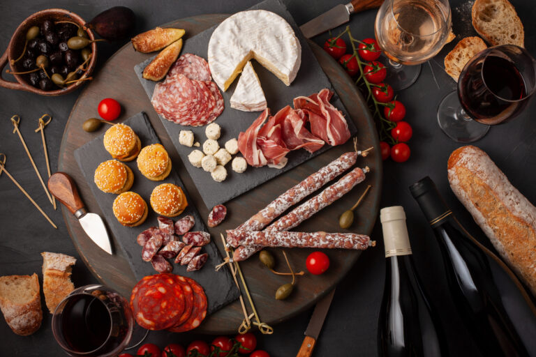 Appetizers table with differents antipasti, cheese, charcuterie, snacks and wine. Mini burgers, sausage, ham, tapas, olives, cheese variety board and baguette over grey concrete background. Top view, flat lay