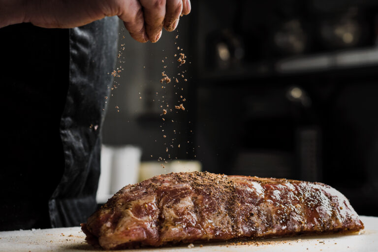 Raw piece of meat, beef ribs. The hand of a male chef puts salt and spices on a dark background, close-up.