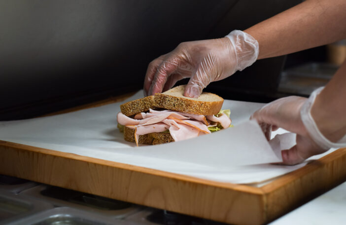 Restaurant kitchen employee wrapping freshly made turkey sandwich for customer order in wax paper