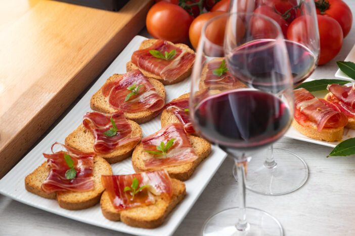 Appetizers of Iberian ham with a glass of wine, ripe tomatoes and cow cheese on a white wooden background.