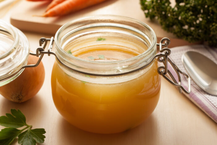 Bone broth made from chicken in a glass jar, with carrots, onions, and celery root in the background