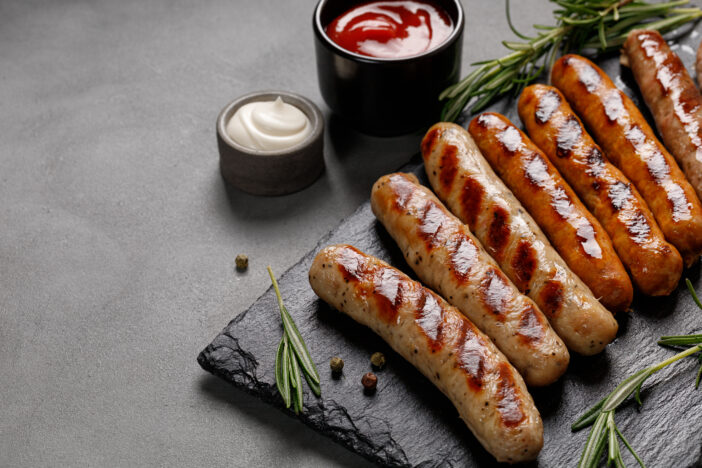 Grilled assorted sausages with sauces and rosemary. Homemade sausages on a stone board. Dark background. Copy space.