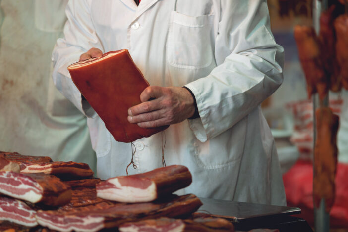 Butcher at Pork Meat Fair with Stacks of Smoked Bacon and other Cured Pork Meat.