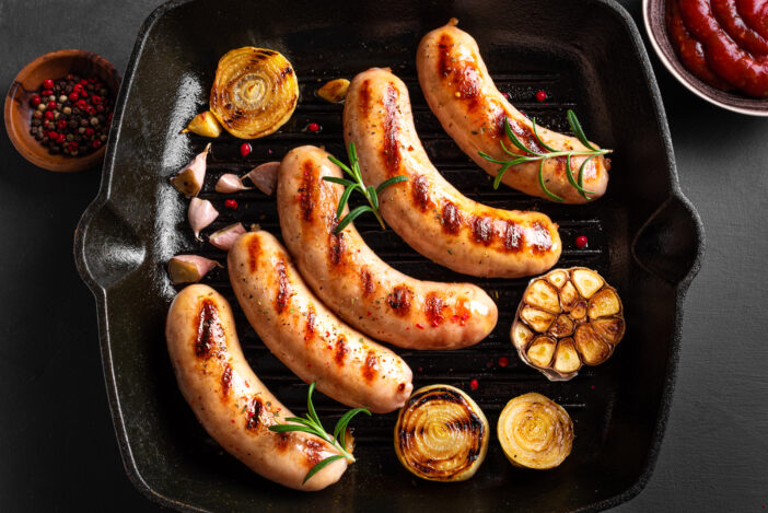 Grilled sausages with vegetables and rosemary on black grill cast iron pan, top view. Roasted homemade wurst sausages and tomato sauce.