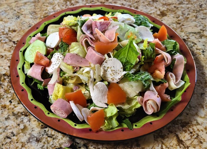 Large and oversized traditional Italian antipasto salad on a large round serving dish which includes Italian meats and cheeses and a variety of vegetables.