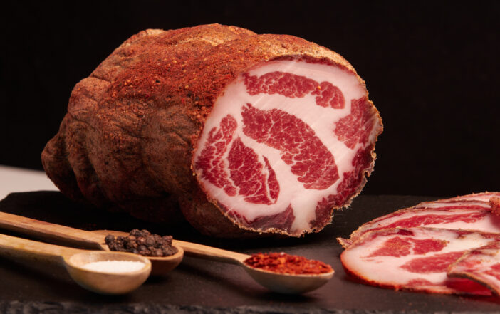 Large piece of cured meat and sliced thin slices of ham with spices on a black serving board. Italian coppa, prosciutto, ham close-up