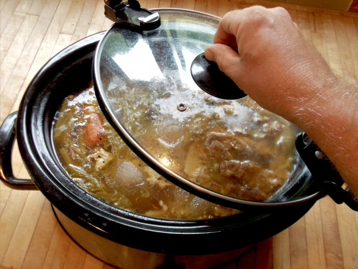 Man lifts up the glass Lid from his Counter Top Crock Pot of Simmering Chicken Soup Broth rich with Fresh Garden Vegetables and Cooking Onions