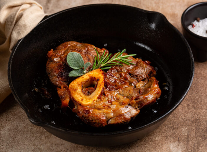 Braised beef steak ossobuco, meat on the bone, cooked with onion, salt, spices, rosemary and sage in an iron cast pan and brown concrete background.