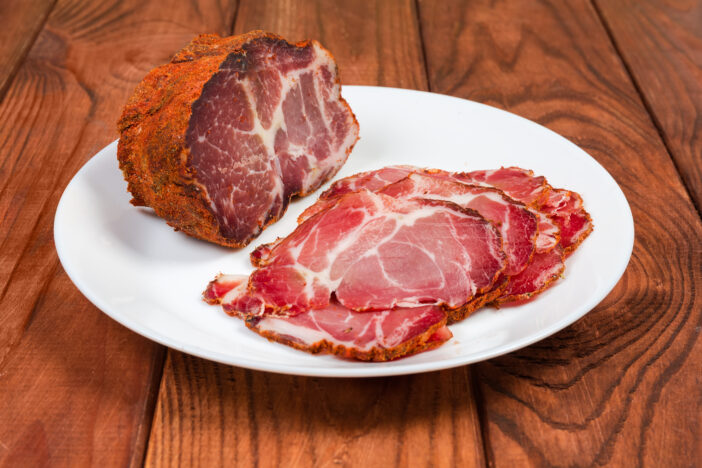 Piece and slices of dried pork neck on the white dish on a dark colored wooden rustic table close-up at selective focus