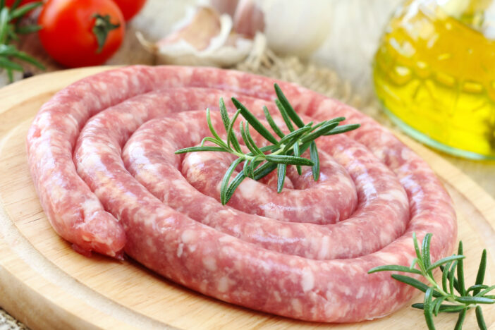 Raw Italian long pork sausage - LUGANEGA - on a wooden cutting board with olive oil and vegetables at the background ready to be cooked