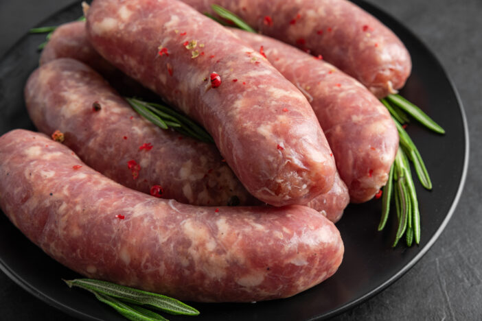 Raw sausages or bratwurst with spices and rosemary in a plate on black background. Close up. Food for grill or barbeque