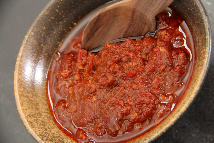 Sambal Oelek, the highly popular condiment of red chili paste. The paste is cooked with oil before being served in a ceramic bowl. A small wooden spoon is tucked in the paste.