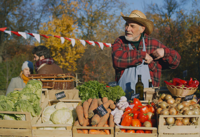 Senior farmer seller stands at the stall with fruits and vegetables, counts cash money. People shopping at local farmers market. Autumn fair. Organic food. Agriculture. Points of sale system.